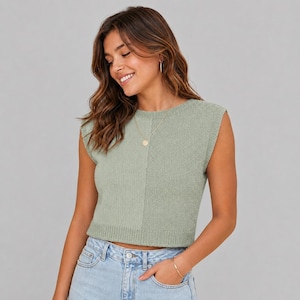 May include: A sage green sleeveless sweater with a round neckline. The sweater is split vertically into two sections. The person is wearing a gold necklace and bracelet, and light blue jeans.