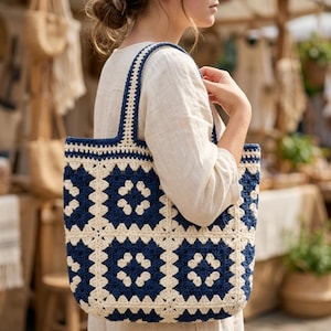 Könnte beinhalten: Handgefertigte Tragetasche mit Granny-Square-Design. Die Tasche hat eine cremefarbene Basis mit marineblauen Quadraten und Trägern. Die Tasche ist aus dickem Garn gefertigt und hat eine robuste Konstruktion, ideal zum Tragen von Gegenständen.