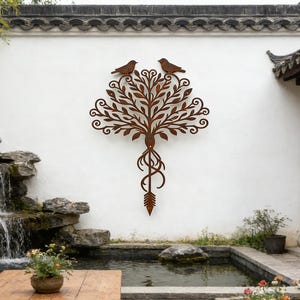 May include: A decorative metal tree wall art with two birds perched on top. The tree is brown with intricate leaf and root details. The art is mounted on a white wall in an outdoor setting with a wooden table and bench.