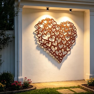 May include: A large, heart-shaped metal wall art piece made up of many smaller hearts. The artwork is a warm, rusty brown colour and is mounted on a white wall, lit by two spotlights. The outdoor setting includes a pathway and greenery.