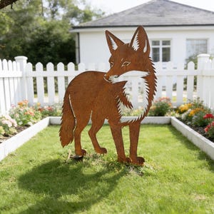 Peut inclure: Sculpture de renard en métal brun rouillé. Le renard est représenté avec des détails, notamment une poitrine et un visage blancs. Il se dresse sur une pelouse verte devant une clôture blanche et une maison.