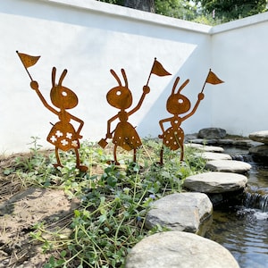 May include: Three rusty metal ant sculptures with flags stand in a garden bed. The ants have cutout dresses and are positioned near a small stream with stepping stones. The background features a white wall and lush greenery.