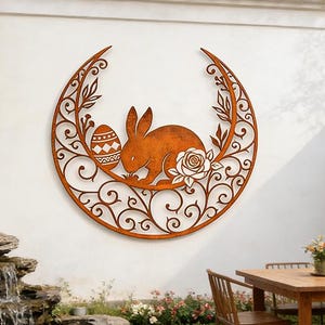 May include: A brown metal wall art piece depicting a rabbit, an Easter egg, and a rose within a crescent moon design. The artwork is mounted on a white wall. The scene includes a garden with a pond and a wooden table. The measurements are in centimeters.