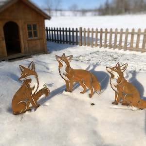 Op de afbeelding: Drie metalen vossensculpturen in een besneeuwde buitenscène. De vossen zijn bruin met witte accenten en staan in verschillende poses. Een klein houten speelhuisje en een hek staan op de achtergrond.