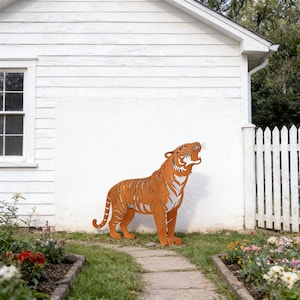 Puede incluir: Una gran escultura de tigre de metal naranja con detalles blancos, rugiendo con la boca abierta. El tigre está sobre hierba frente a una pared blanca y una valla blanca. La escultura es un elemento decorativo de jardín.