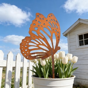 Pode incluir: Uma estaca de jardim de metal laranja enferrujado decorativa em forma de borboleta. A borboleta tem um design de asa detalhado com padrões recortados. É colocada num vaso branco com tulipas brancas, contra uma cerca branca e um pequeno edifício branco.