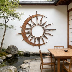 Puede incluir: Una gran pieza de arte mural de metal oxidado que representa un diseño de sol y media luna. La obra de arte está montada en una pared blanca en un entorno al aire libre. El sol tiene rayos puntiagudos y la luna se curva alrededor del sol.