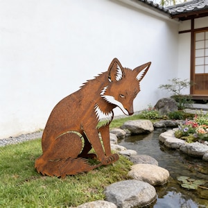 May include: A weathered brown metal fox sculpture in a seated pose, with white detailing on the chest and ears. The fox is positioned on green grass beside a small pond with rocks, in an outdoor setting.
