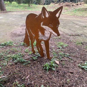 Peut inclure: Une sculpture de jardin en métal brun rouillé représentant un renard. Le renard est représenté en position de marche avec des détails, notamment des oreilles pointues et une queue touffue. La sculpture est placée dans un jardin.