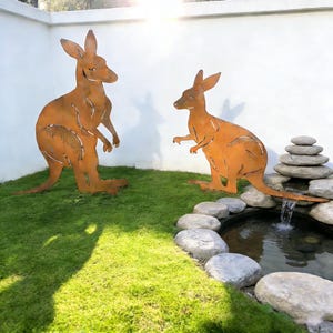 Puede incluir: Dos grandes esculturas de canguros de metal naranja oxidado sobre césped verde. Los canguros son siluetas recortadas con detalles. Un pequeño estanque con cascada y piedras apiladas se encuentra en el fondo.