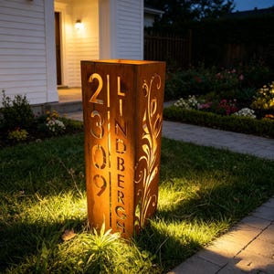 Pode incluir: Uma placa de endereço retangular de metal desgastado com os números 21309 e o nome da rua LINDBERGH recortados. A placa tem um design floral em um lado e é iluminada por dentro, criando um brilho quente. É colocada em um jardim da frente.