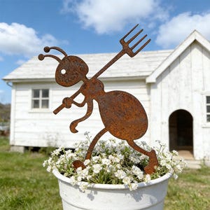 Könnte beinhalten: Ein rostiges Metall-Ameisen-Gartenstecker mit einer Gabel über der Schulter. Die Ameise befindet sich in einem weißen Blumentopf mit kleinen weißen Blumen. Im Hintergrund befindet sich ein weißes Haus.