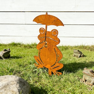 May include: A rusty metal frog sculpture holding an umbrella, standing on green grass. The frog has cut-out eyes and a smiling mouth. Several other frog statues are in the background, creating a whimsical garden scene.