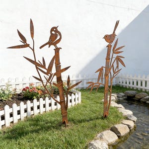 Può includere: Due sculture di uccelli in metallo arrugginito appollaiate su steli di bambù con foglie. Le sculture sono collocate in un giardino con una staccionata bianca, erba e un piccolo ruscello. Gli uccelli sono rivolti in direzioni diverse.