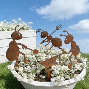 May include: Three rusty metal ant garden ornaments in a white flower pot filled with white flowers. One ant holds a shovel, another a pitchfork, and the third pushes a wheelbarrow-like tool. The background is a blue sky with clouds.