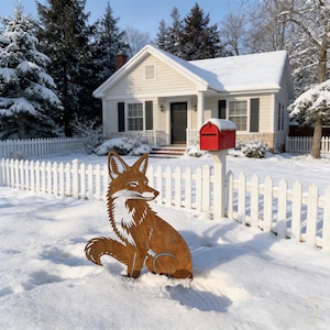 May include: A metal fox yard decoration sits in the snow in front of a white picket fence and a house. The fox is brown with white accents and is in a seated position. A red mailbox is also visible.