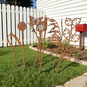 May include: A display of rusty metal garden sculptures depicting a butterfly, seed heads, and other plant forms, set in a grassy garden. A white picket fence and a red postbox are in the background, creating a rustic aesthetic.