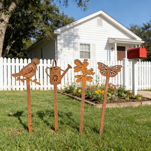 May include: Four rusty metal garden stakes with nature-themed cutouts: a bird on a branch, a watering can, a fairy, and a butterfly. The stakes are placed in a grassy yard in front of a white picket fence and a house.