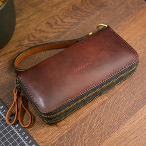May include: A rectangular dark brown leather clutch wallet with a wrist strap, featuring two brass-coloured metal zippers and the embossed text SAVOROV on the front. The wallet is positioned next to a black measuring tool with white markings.