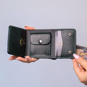 Peut inclure: Un portefeuille trifold en cuir vert foncé avec des coutures vertes apparentes, une poche centrale pour monnaie à bouton-pression et plusieurs fentes pour cartes. Le mot FAVOROV est embossé sur la poche, et deux boutons-pression argentés sont situés sur le côté droit.