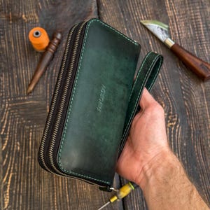 May include: A dark green leather clutch wallet with two zippered compartments and a matching wrist strap. The front of the wallet features the embossed text FAVOROV. The wallet is constructed with visible green stitching along the edges.