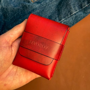 Puede incluir: Un tarjetero de cuero rojo brillante que presenta una correa horizontal en la parte frontal con el texto FAVOROV grabado. El accesorio tiene un acabado liso con bordes de color rojo más oscuro y una forma rectangular estructurada.