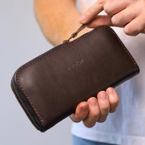 May include: A dark brown leather zip-around wallet featuring a smooth finish and visible stitching along the edges. The word FAVOROV is embossed in the center of the front panel. The wallet includes a metal zipper pull and a dark zipper track.