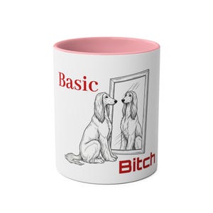 Puede incluir: Taza de cerámica blanca con interior rosa. La taza presenta una ilustración en blanco y negro de un perro mirándose en un espejo. Las palabras "Basic" y "Bitch" están impresas en rojo.