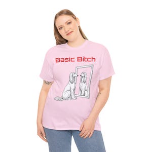 May include: A light pink t-shirt featuring the red text "Basic Bitch" above a black and white line drawing of a long-haired dog sitting in front of a rectangular mirror, with the dog's reflection visible in the glass surface.