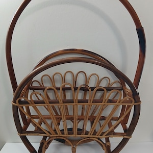 Puede incluir: Un revistero redondo vintage de ratán con asa. El revistero presenta un diseño de cesta tejida con un patrón en forma de abanico y un acabado marrón oscuro. El asa es de madera curvada, marrón oscuro.