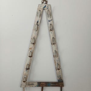 Puede incluir: Un estante de pared triangular de metal vintage con un acabado blanco desgastado y detalles en azul. El estante presenta múltiples ranuras y ganchos, y se suspende mediante un gancho dorado en la parte superior. La pieza muestra signos de edad y desgaste.