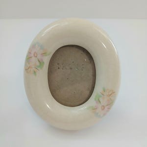 May include: An oval-shaped ceramic picture frame with a cream-colored crackle glaze finish. The frame features delicate hand-painted floral motifs in soft shades of pink, yellow, and green on the left and right sides, surrounding a central oval opening.