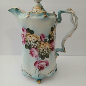 May include: A vintage porcelain pitcher with a light blue and white background. The pitcher features a floral design with pink roses and gold accents. The handle and rim are also trimmed in gold.