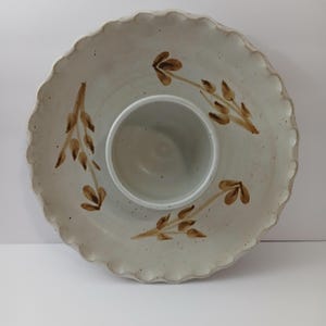 Puede incluir: Una fuente de cerámica con un cuenco central para salsas. La fuente de color blanco roto tiene un borde festoneado y está decorada con diseños marrones en forma de hojas. La fuente es adecuada para servir aperitivos.