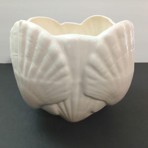 May include: A glossy off-white ceramic planter or bowl featuring a molded exterior design of multiple overlapping scallop shells. The vessel has a wide, open top and a rounded base, with the shell textures creating a repeating pattern around the sides.