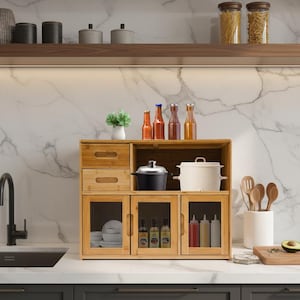 May include: A bamboo kitchen cabinet with glass doors and drawers. The cabinet contains kitchen items such as pots, bottles, and condiments. A small plant and wooden utensils are also visible.