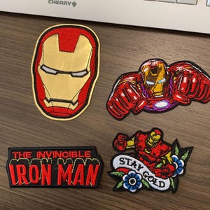 May include: Four embroidered patches featuring Iron Man designs. One patch is the Iron Man mask in gold and red. Another shows Iron Man in a fighting pose. A third patch reads "THE INVINCIBLE IRON MAN". The last patch shows Iron Man with a "Stay Gold" banner.