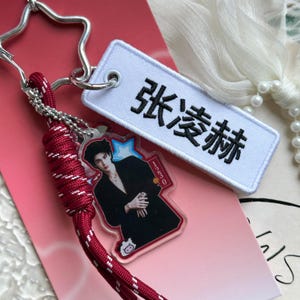 May include: A keychain featuring a star-shaped metal ring, a red paracord lanyard with white flecks, and a rectangular white embroidered patch with black Chinese characters. An acrylic charm displays a person in a black suit with the text 1230.