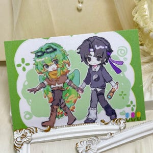 Könnte beinhalten: Eine rechteckige Karte mit einer farbenfrohen Illustration zweier Anime-Figuren, die Händchen halten. Eine Figur hat grünes Haar und eine Schutzbrille, die andere dunkles Haar, ein violettes Stirnband und ein Yin-Yang-Symbol auf dem Shirt.