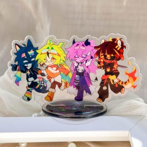May include: An acrylic standee featuring four stylised cartoon characters in a row. The characters have distinct colour palettes including blue with rainbow accents, yellow and orange, pink and purple, and red and black with flame-like details.