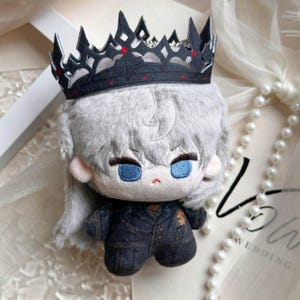May include: A plush doll featuring light gray hair, bright blue embroidered eyes, and a small red mouth. The doll wears a black crown with intricate cutouts and red accents, paired with a dark charcoal gray jacket with subtle gold detailing.