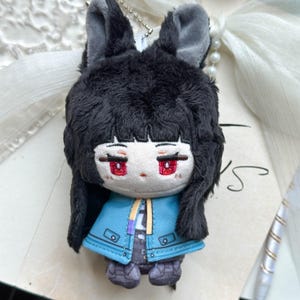 May include: A plush doll featuring black fur, tall ears, and a pale face with embroidered red eyes. The doll wears a bright blue jacket with yellow and purple accents over a dark patterned outfit. A silver ball chain is attached to the top of the head.