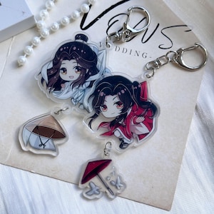 Puede incluir: Dos llaveros de acrílico con personajes de anime estilo chibi de cabello oscuro. Un personaje viste túnicas blancas con un dije de sombrero cónico, y el otro viste túnicas rojas con un dije de paraguas rojo. Ambos tienen cierres de mosquetón.