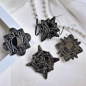 May include: Four metal enamel pins featuring intricate black and silver geometric designs. The pins display celestial motifs including a crescent moon, a sunburst, and complex star-like patterns with sharp points and detailed linework.