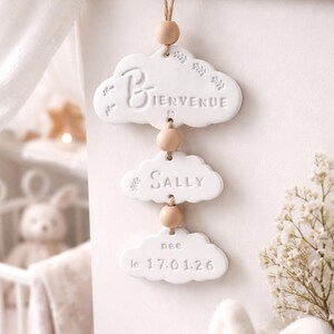 Op de afbeelding: Een witte, wolk-vormige wanddecoratie met het woord "Bienvenue" en de naam "Sally". De onderste wolk leest "nee le 17.01.26". De wolken zijn verbonden met houten kralen en touw.