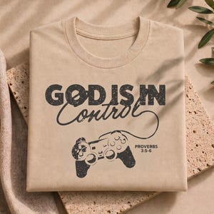 Puede incluir: Una camiseta beige con un gráfico negro desgastado que dice "GOD IS IN CONTROL" sobre una ilustración de un mando de videojuegos. El cable del mando forma un bucle y el texto "PROVERBS 3:5-6" aparece impreso a la derecha.