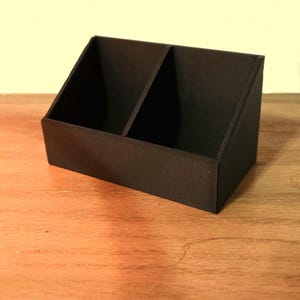 Puede incluir: Un organizador de escritorio negro de dos compartimentos con la parte trasera inclinada. El organizador está hecho de un material sólido y se asienta sobre una superficie de madera. Los compartimentos están divididos por un divisor vertical.