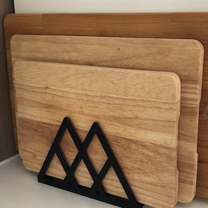 May include: Wooden cutting boards in various sizes, displayed in a black metal mountain-shaped holder. The boards have a natural wood grain finish. A larger wooden board is visible in the background. The holder adds a decorative touch to the kitchen.