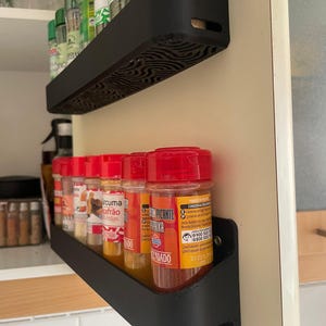 Könnte beinhalten: Schwarzes, an der Wand befestigtes Gewürzregal mit zwei Ebenen, gefüllt mit Gewürzgläsern. Die Gläser haben rote Deckel und verschiedene Etiketten. Das Regal ist an einer cremefarbenen Wand befestigt. Die Regale sind aus schwarzem Material.
