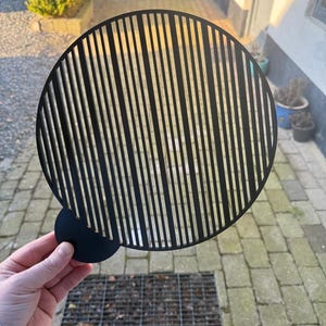 Puede incluir: Una pieza decorativa de metal negro con un diseño circular con líneas verticales. Un círculo negro más pequeño y sólido está unido al círculo más grande. La pieza se sostiene contra un fondo de un patio de ladrillos.